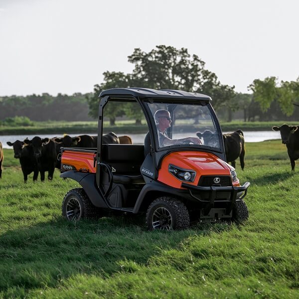 Une vue d'un conducteur au volant d'un véhicule utilitaire orange Kubota RTV transportant des matériaux légers dans la benne arrière, traversant un terrain agricole ensoleillé.
